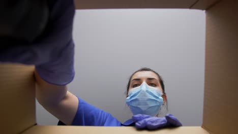 Woman-in-Mask-Packing-Camera-Into-Parcel-Box.delivery,-shipping-and-pandemic-concept-woman-in-protective-medical-mask-and-gloves-packing-digital-camera-and-lens-into-parcel-box