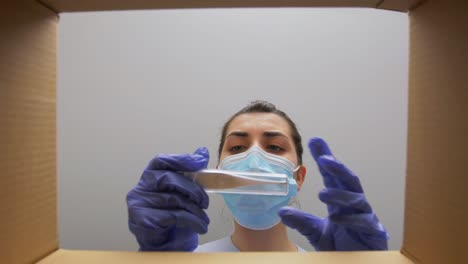 Mujer-Con-Mascarilla-Abriendo-Caja-De-Paquete-Con-Ropa.-Entrega-A-Domicilio,-Envío-Y-Concepto-De-Pandemia.-Mujer-Con-Mascarilla-Médica-Protectora-Y-Guantes-Abriendo-Caja-De-Paquete,-Mirando-Dentro-Y-Sacando-Ropa.