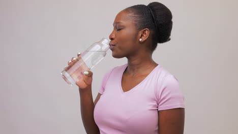 Mujer-Africana-Feliz-Bebiendo-Agua-De-Una-Botella-De-Vidrio.-Retrato-De-Una-Joven-Afroamericana-Feliz-Y-Sonriente-Bebiendo-Agua-De-Una-Botella-De-Vidrio-Reutilizable-Sobre-Fondo-Gris