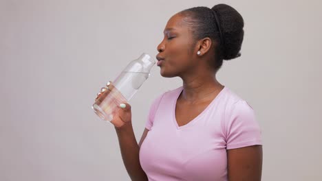 Mujer-Africana-Feliz-Bebiendo-Agua-De-Una-Botella-De-Vidrio.-Retrato-De-Una-Joven-Afroamericana-Feliz-Y-Sonriente-Bebiendo-Agua-De-Una-Botella-De-Vidrio-Reutilizable-Sobre-Fondo-Gris