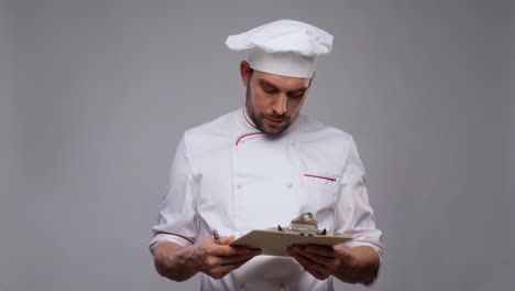 Concepto-De-Cocina,-Culinaria-Y-Personas.-Chef-Masculino-Con-Portapapeles-Y-Bolígrafo-Escribiendo-Sobre-Fondo-Gris.