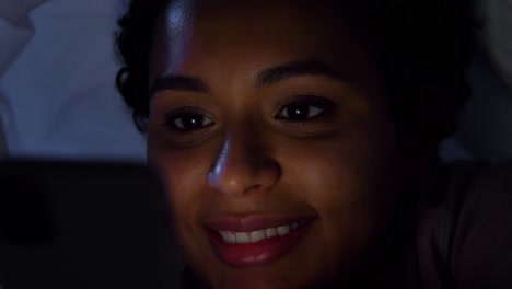 Woman-with-Smartphone-Under-Duvet-in-Bed-at-Night.technology,-internet,-communication-and-people-concept-happy-smiling-young-african-american-woman-with-smartphone-lying-under-blanket-in-bed-at-home-at-night