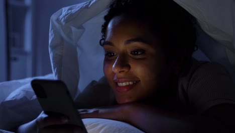 Frau-Mit-Smartphone-Unter-Der-Bettdecke-Im-Bett-In-Der-Nacht.Technologie,-Internet,-Kommunikation-Und-Menschen-Konzept-Glücklich-Lächelnde-Junge-Afroamerikanische-Frau-Mit-Smartphone,-Die-Nachts-Zu-Hause-Unter-Der-Decke-Im-Bett-Liegt