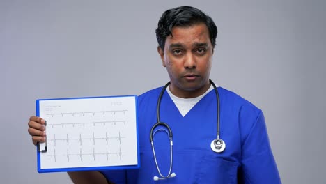 Doctor-with-Cardiogram-Having-Video-Conference.healthcare,-medicine-and-communication-concept-happy-smiling-indian-doctor-or-male-nurse-in-blue-uniform-with-cardiogram-on-clipboard-having-video-conference-over-grey-background