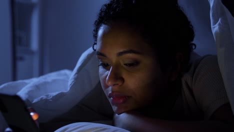 Woman-with-Smartphone-in-Bed-at-Home-at-Night.technology,-internet,-communication-and-people-concept-young-african-american-woman-with-smartphone-lying-in-bed-at-home-at-night