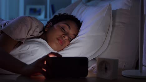 Mujer-Dormida-Que-Se-Despierta-Por-El-Teléfono-Por-La-Noche.-Tecnología,-Internet,-Comunicación-Y-Concepto-De-Personas.-Mujer-Afroamericana-Joven-Durmiendo-En-La-Cama-Y-Despertándose-Por-El-Teléfono-Inteligente-En-Casa-Por-La-Noche.