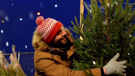 Hombre-Feliz-Comprando-Un-árbol-De-Navidad-En-El-Mercado.-Vacaciones-De-Invierno-Y-Concepto-De-Personas:-Hombre-Feliz-Y-Sonriente-Comprando-Un-árbol-De-Navidad-En-El-Mercado-Callejero-Por-La-Noche-Mientras-Cae-La-Nieve