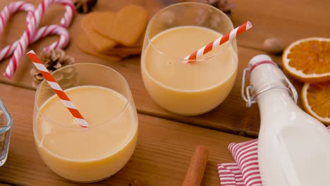 Glasses-of-Eggnog,-Ingredients-and-Spices-on-Wood.christmas,-winter-holidays-and-seasonal-drinks-concept-–-glasses-with-eggnog,-ingredients-and-aromatic-spices-on-wooden-board-over-snow-falling