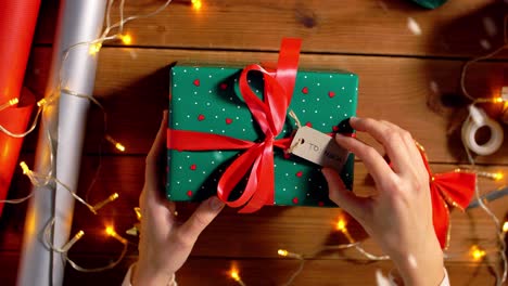 Woman-Packing-Christmas-Gifts-at-Home-Over-Snow.holidays,-new-year-and-christmas-concept-–-woman-holding-gift-box-in-green-wrapping-paper-package-with-red-bow-and-name-tag-at-home-over-snow