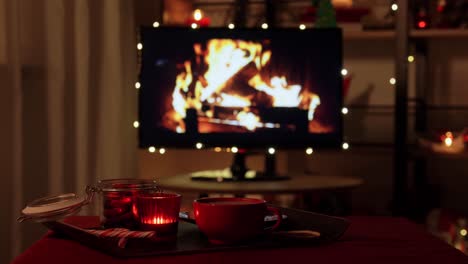 Weihnachtsleckereien-Und-Getränke-Auf-Dem-Tisch-Im-Gemütlichen-Zuhause.-Winterferien-Und-Hygge-Konzept-–-Hand-Stellt-Eine-Tasse-Kaffee-Oder-Heiße-Schokolade-Mit-Ingwerkeksen,-Kerze,-Zuckerstangen-Und-Geschenkboxen-Auf-Den-Tisch-über-Einem-Fernsehmonitor,-Der-Als-Kamin-Im-Gemütlichen-Zuhause-Genutzt-Wird