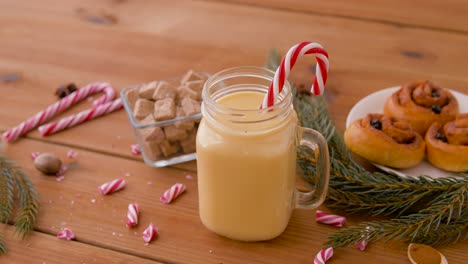 Glass-Mug-of-Eggnog,-Ingredients-and-Sweets.christmas,-winter-holidays-and-seasonal-drinks-concept-–-glass-mug-with-eggnog-and-candy-cane-decoration,-sweets-and-aromatic-spices-on-wooden-background-over-snow-falling