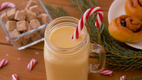 Glass-Mug-of-Eggnog,-Ingredients-and-Sweets.christmas,-winter-holidays-and-seasonal-drinks-concept-–-glass-mug-with-eggnog-and-candy-cane-decoration,-sweets-and-aromatic-spices-on-wooden-background-over-snow-falling