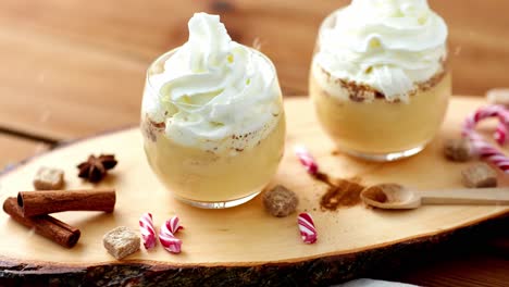 Glasses-of-Eggnog,-Candy-Canes-and-Spices-on-Wood.christmas-and-seasonal-drinks-concept-–-snow-falling-over-glasses-of-eggnog-with-whipped-cream-topping,-crashed-candy-canes-and-spices-on-wooden-background