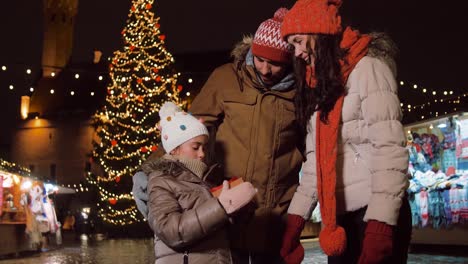 Familia-Feliz-Con-Regalo-En-El-Mercado-Navideño-De-La-Ciudad.-Concepto-De-Celebración-Y-Vacaciones-De-Invierno:-Familia-Feliz-Con-Regalo-En-El-Mercado-Navideño-En-La-Plaza-Del-Ayuntamiento-De-Tallin,-Estonia