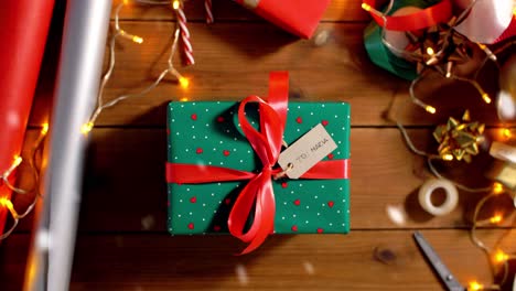 Woman-Packing-Christmas-Gifts-at-Home-Over-Snow.holidays,-new-year-and-christmas-concept-–-woman-holding-gift-box-in-green-wrapping-paper-package-with-red-bow-and-name-tag-at-home-over-snow