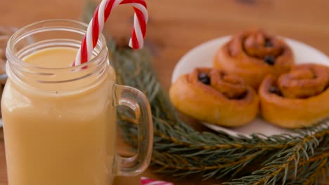 Glass-Mug-of-Eggnog,-Ingredients-and-Sweets.christmas,-winter-holidays-and-seasonal-drinks-concept-–-glass-mug-with-eggnog-and-candy-cane-decoration,-sweets-and-aromatic-spices-on-wooden-background-over-snow-falling