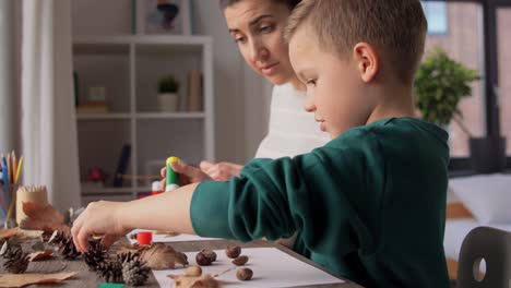 Mother-and-Son-Making-Pictures-of-Autumn-Leaves.family,-creativity-and-craft-concept-–-mother-and-little-son-with-glue-sticks-and-paper-making-pictures-of-dry-autumn-leaves,-pine-cones-and-chestnuts-at-home