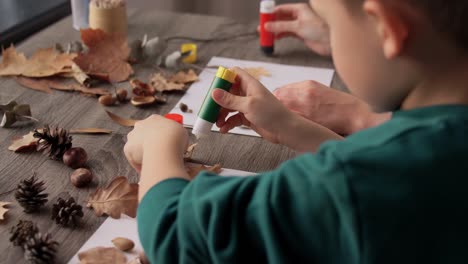 Mother-and-Son-Making-Pictures-of-Autumn-Leaves.family,-creativity-and-craft-concept-–-mother-and-little-son-with-glue-sticks-and-paper-making-pictures-of-dry-autumn-leaves,-pine-cones-and-chestnuts-at-home