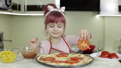 Cooking-pizza.-Little-child-in-apron-adding-sliced-tomatoes-to-dough-in-kitchen