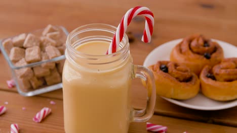 Glass-Mug-of-Eggnog,-Ingredients-and-Sweets.christmas,-winter-holidays-and-seasonal-drinks-concept-–-glass-mug-with-eggnog-and-candy-cane-decoration,-sweets-and-aromatic-spices-on-wooden-background-over-snow-falling