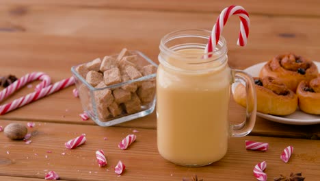 Glass-Mug-of-Eggnog,-Ingredients-and-Sweets.christmas,-winter-holidays-and-seasonal-drinks-concept-–-glass-mug-with-eggnog-and-candy-cane-decoration,-sweets-and-aromatic-spices-on-wooden-background-over-snow-falling