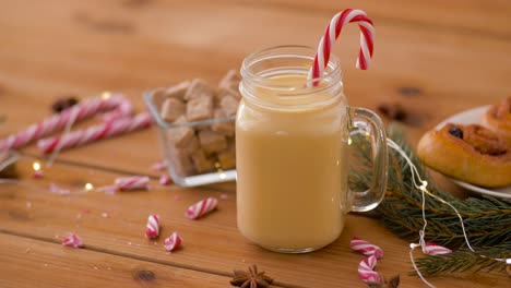 Glass-Mug-of-Eggnog,-Ingredients-and-Sweets.christmas,-winter-holidays-and-seasonal-drinks-concept-–-glass-mug-with-eggnog-and-candy-cane-decoration,-sweets-and-aromatic-spices-on-wooden-background-over-snow-falling