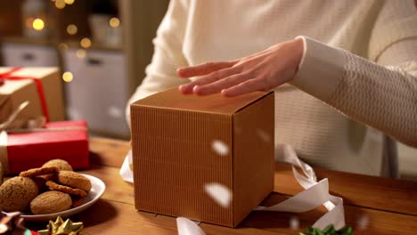 Mujer-Abriendo-Un-Regalo-De-Navidad-En-Casa-Sobre-La-Nieve.-Vacaciones,-Año-Nuevo-Y-Concepto-De-Celebración:-Mujer-Sentada-A-La-Mesa-Y-Abriendo-Un-Regalo-De-Navidad-En-Casa-Sobre-La-Nieve