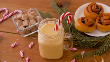 Glass-Mug-of-Eggnog,-Ingredients-and-Sweets.christmas,-winter-holidays-and-seasonal-drinks-concept-–-glass-mug-with-eggnog-and-candy-cane-decoration,-sweets-and-aromatic-spices-on-wooden-background-over-snow-falling