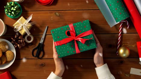 Woman-Packing-Christmas-Gifts-at-Home-Over-Snow.holidays,-new-year-and-christmas-concept-–-woman-holding-gift-box-in-green-wrapping-paper-package-with-red-bow-and-name-tag-at-home-over-snow