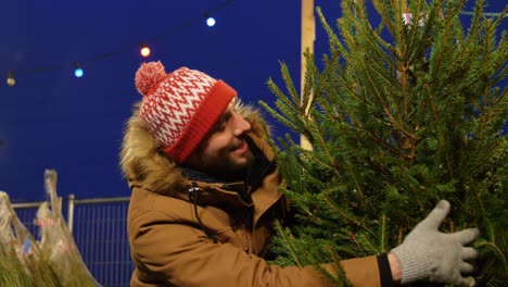 Hombre-Feliz-Comprando-Un-árbol-De-Navidad-En-El-Mercado.-Vacaciones-De-Invierno-Y-Concepto-De-Personas:-Hombre-Feliz-Y-Sonriente-Comprando-Un-árbol-De-Navidad-En-El-Mercado-Callejero-Por-La-Noche-Mientras-Cae-La-Nieve