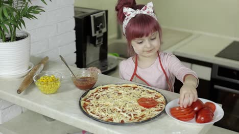 Cooking-pizza.-Little-child-in-apron-adding-sliced-tomatoes-to-dough-in-kitchen