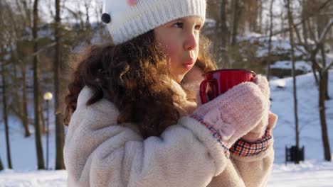 Niña-Con-Una-Taza-De-Té-Caliente-En-El-Parque-De-Invierno.-Concepto-De-Infancia,-Ocio-Y-Temporada:-Niña-Feliz-Con-Una-Taza-De-Té-Caliente-En-El-Parque-De-Invierno-Mientras-Cae-La-Nieve