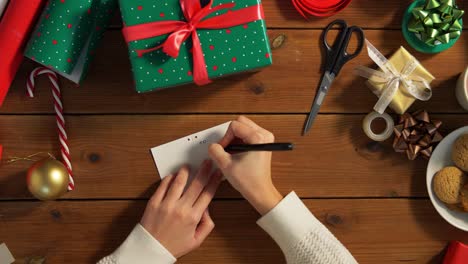 Hands-Attaching-Name-Tag-To-Christmas-Gift.holidays,-new-year-and-christmas-concept-–-hands-packing-gift-box,-attaching-tag-and-writing-receiver-name-at-home-over-snow