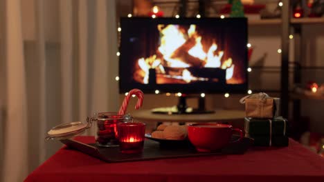 Weihnachtsleckereien-Und-Getränke-Auf-Dem-Tisch-Im-Gemütlichen-Zuhause.-Winterferien-Und-Hygge-Konzept-–-Hand-Stellt-Eine-Tasse-Kaffee-Oder-Heiße-Schokolade-Mit-Ingwerkeksen,-Kerze,-Zuckerstangen-Und-Geschenkboxen-Auf-Den-Tisch-über-Einem-Fernsehmonitor,-Der-Als-Kamin-Im-Gemütlichen-Zuhause-Genutzt-Wird