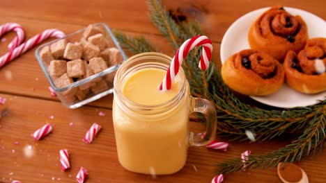 Glass-Mug-of-Eggnog,-Ingredients-and-Sweets.christmas,-winter-holidays-and-seasonal-drinks-concept-–-glass-mug-with-eggnog-and-candy-cane-decoration,-sweets-and-aromatic-spices-on-wooden-background-over-snow-falling