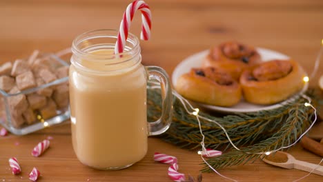 Glass-Mug-of-Eggnog,-Ingredients-and-Sweets.christmas,-winter-holidays-and-seasonal-drinks-concept-–-glass-mug-with-eggnog-and-candy-cane-decoration,-sweets-and-aromatic-spices-on-wooden-background-over-snow-falling