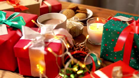 Weihnachtsgeschenke,-Kerzen,-Kaffee-Und-Lichterketten.-Feiertage,-Neujahr-Und-Weihnachtskonzept-–-Geschenkboxen-Mit-Namensschildern,-Kerze,-Heißem-Schokoladengetränk-Im-Becher-Und-Elektrischen-Lichterketten-Auf-Einem-Holztisch-über-Schnee