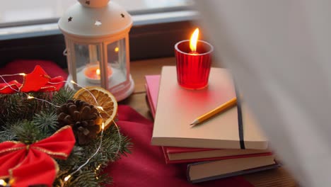 Christmas-Wreath-Books-Candle-Lantern-on-Window.winter-holidays,-new-year-and-decorations-concept-–-christmas-fir-wreath,-books,-candle-and-lantern-on-window-sill-at-home-over-snow