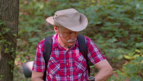 Senior-old-tourist-man-lost-and-looking-at-compass,-examining-route-while-having-walk-in-summer-wood