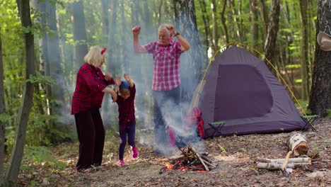 Senior-elderly-grandmother-grandfather-with-granddaughter-dancing-celebrating-over-campfire-in-wood.