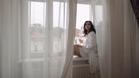 Bride-in-boudoir-dress-sitting-on-window-sill-wedding-morning-preparations-woman-in-night-gown,-veil