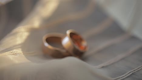 Two-gold-wedding-rings-lying-on-white-gray-surface-of-dress-shining-with-light-close-up-macro