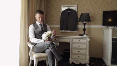 Handsome-groom-man-with-wedding-bouquet-in-his-hands-at-home-near-window-preparing-to-go-to-bride