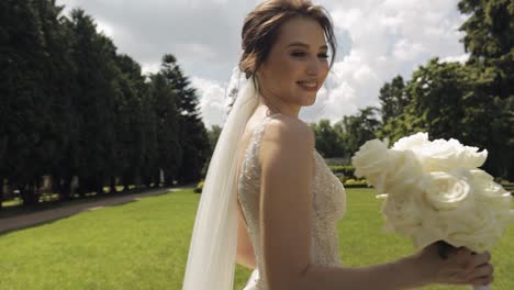 Beautiful-stylish-bride-in-white-wedding-dress-and-veil-holding-wedding-bouquet-in-hands-in-park