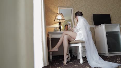 Bride-in-white-boudoir-dress-sitting-near-mirror-looking-at-reflection,-wedding-preparations-at-home