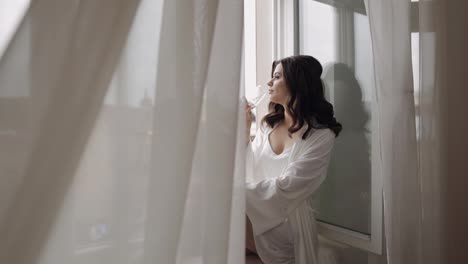 Bride-in-boudoir-dress-sitting-on-window-sill-wedding-morning-preparations-woman-in-night-gown,-veil