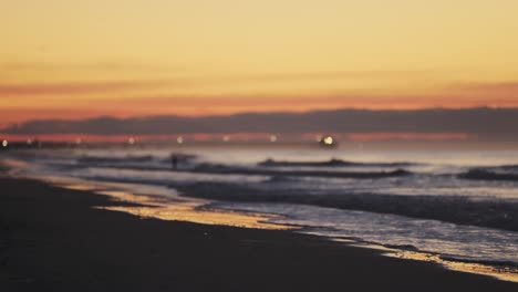 Toma-Panorámica-De-Una-Hermosa-Playa-De-Arena-Temprano-En-La-Mañana-Y-El-Océano-Borroso-Y-El-Muelle-Del-Embarcadero-Iluminado-En-El-Fondo