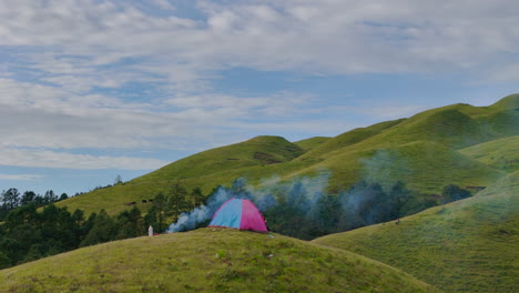 Drone-shot-reveals-mesmerizing-beauty-of-a-peaceful-hill-landscape-in-Sailung,-Dolakha,-Nepal-male-camper-enjoys-a-bonfire-beside-his-tent-under-a-blue-sky,-highlighting-a-heavenly-camping-experience