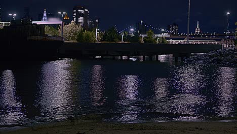 Puente-De-Brooklyn,-De-Noche,-Junto-Al-Agua,-Filmado-Desde-Una-Playa-De-Arena,-Cerca-De-Un-Muelle