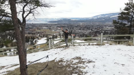 Vista-Aérea,-Joven-Caminando-Sobre-Una-Cuerda-En-Un-Paisaje-Nevado-De-Invierno-Sobre-Kelowna,-Columbia-Británica,-Canadá,-Fotografía-Tomada-Con-Un-Dron-En-órbita